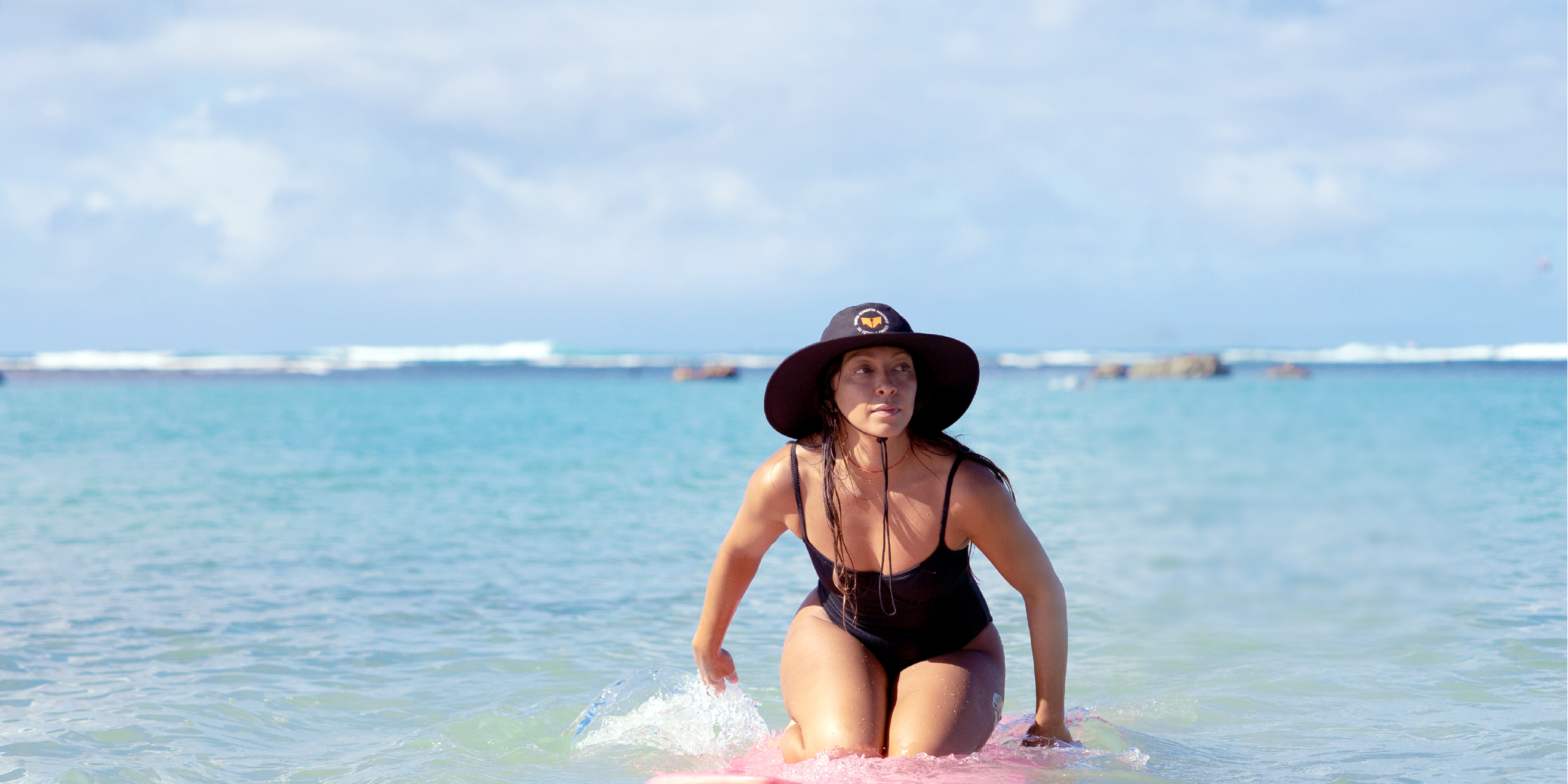 Woman kneeling on a surfboard paddles in the ocean wearing a wide brim black Vertra hat.